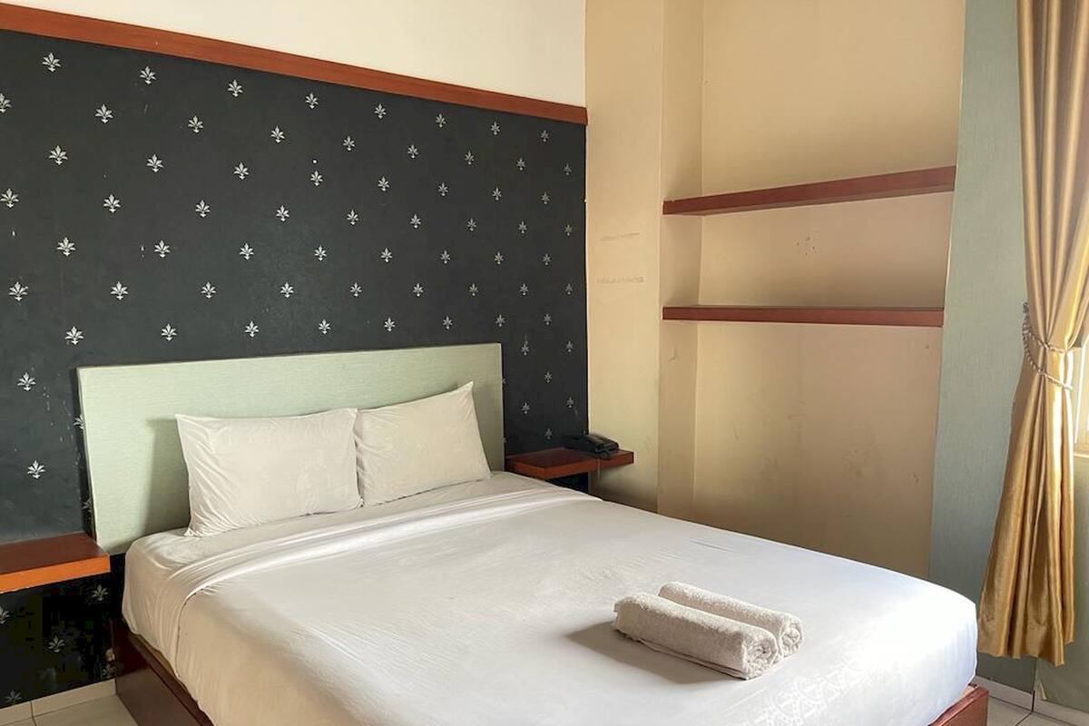 Een net opgemaakt bed met witte lakens en een opgerolde handdoek, een nachtkastje met een telefoon, en een muur met een bloemenprint.