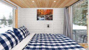 Une chambre à coucher avec une couvre-lit à carreaux, un tableau représentant une scène de forêt accroché au mur, et une fenêtre donnant sur les arbres.