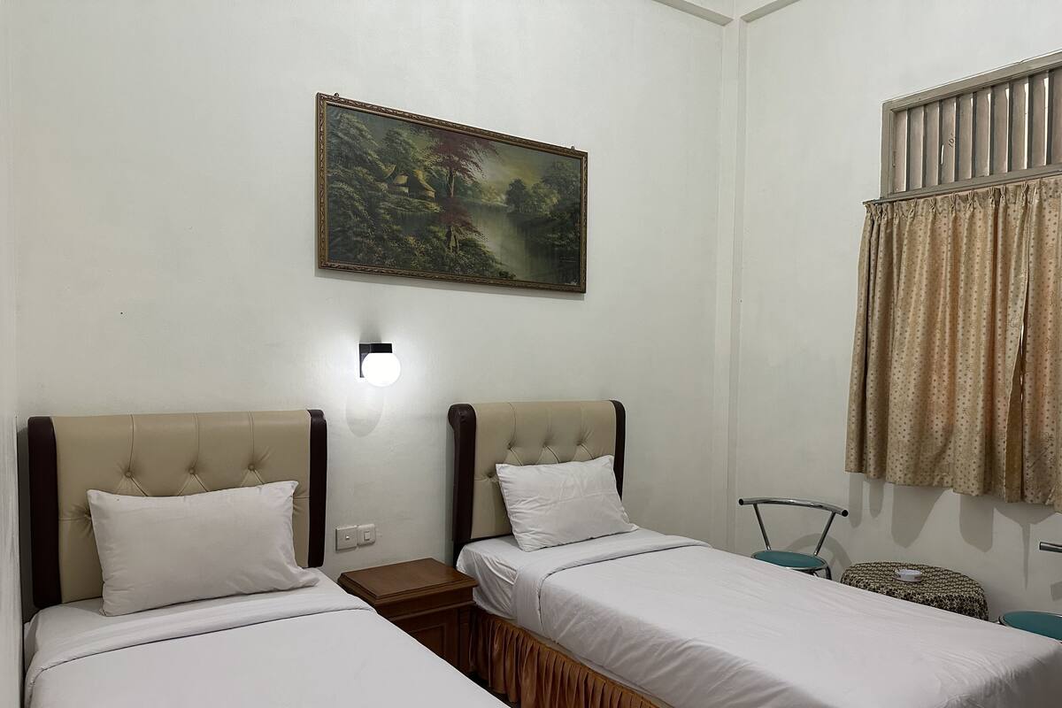 Een hotelkamer met twee eenpersoonsbedden, een nachtkastje, een wandlamp, een schilderij en een raam met gordijnen.