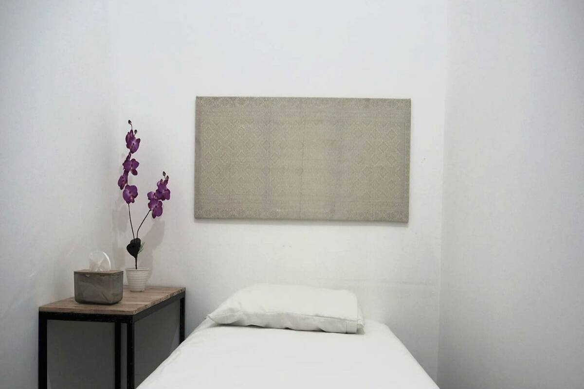 A minimalist bedroom with a single bed, a nightstand, a vase with purple flowers, and a plain wall.