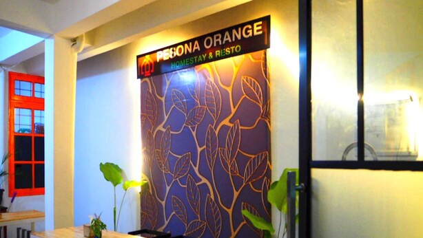 A reception area with a sign reading "PESONA ORANGE HOMESTAY & RESTO". There is a reception desk with chairs, a plant, and a calendar on the wall.
