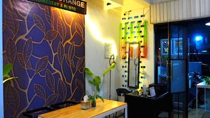 A modern interior with a sign reading "PESONA ORANGE HOMESTAY & RESTO". There is a wooden table with chairs, a potted plant, and a wall with a leaf pattern.