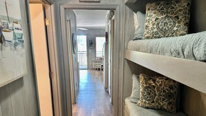 Un couloir étroit au plancher en bois, avec des lits superposés d’un côté et une porte donnant sur une autre pièce.