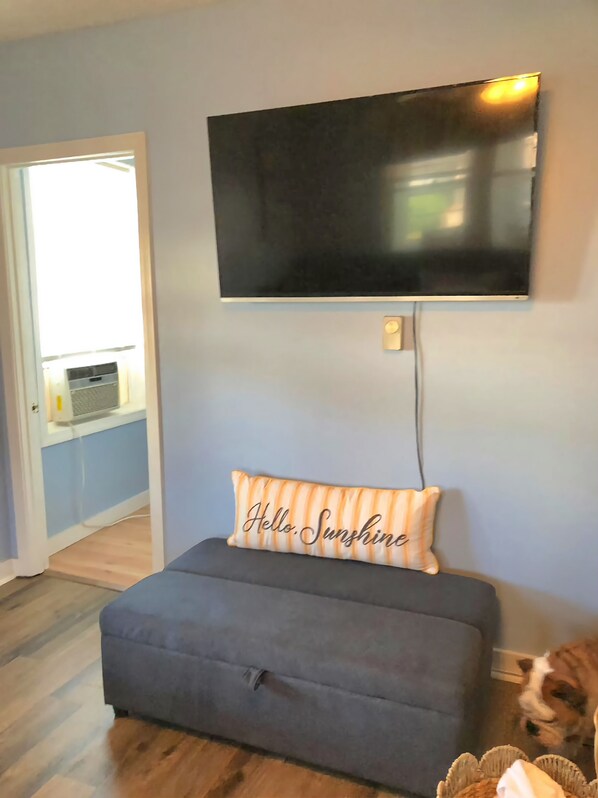 Una habitación con un sofá gris, un televisor montado en la pared y una almohada con la inscripción 'Hello. Sunshine'.