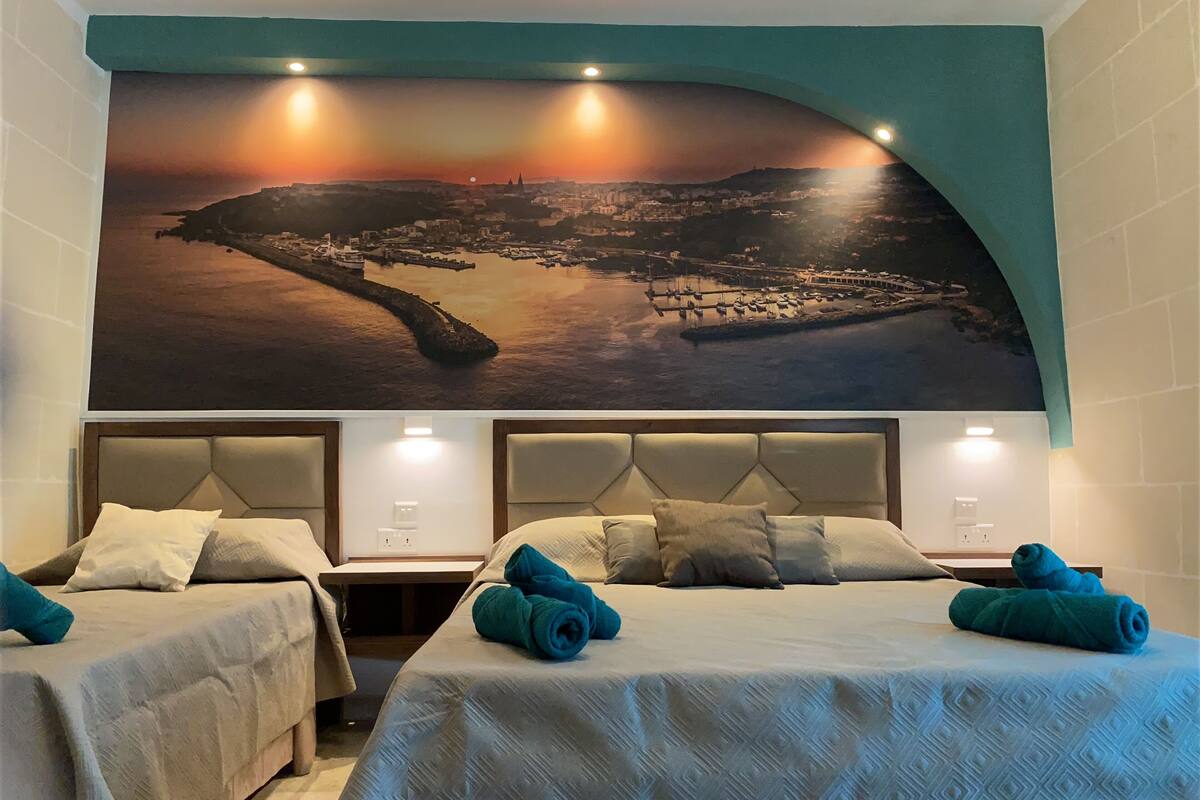 Habitación de hotel con dos camas, un mural grande con un paisaje urbano en la pared y vista al mar.