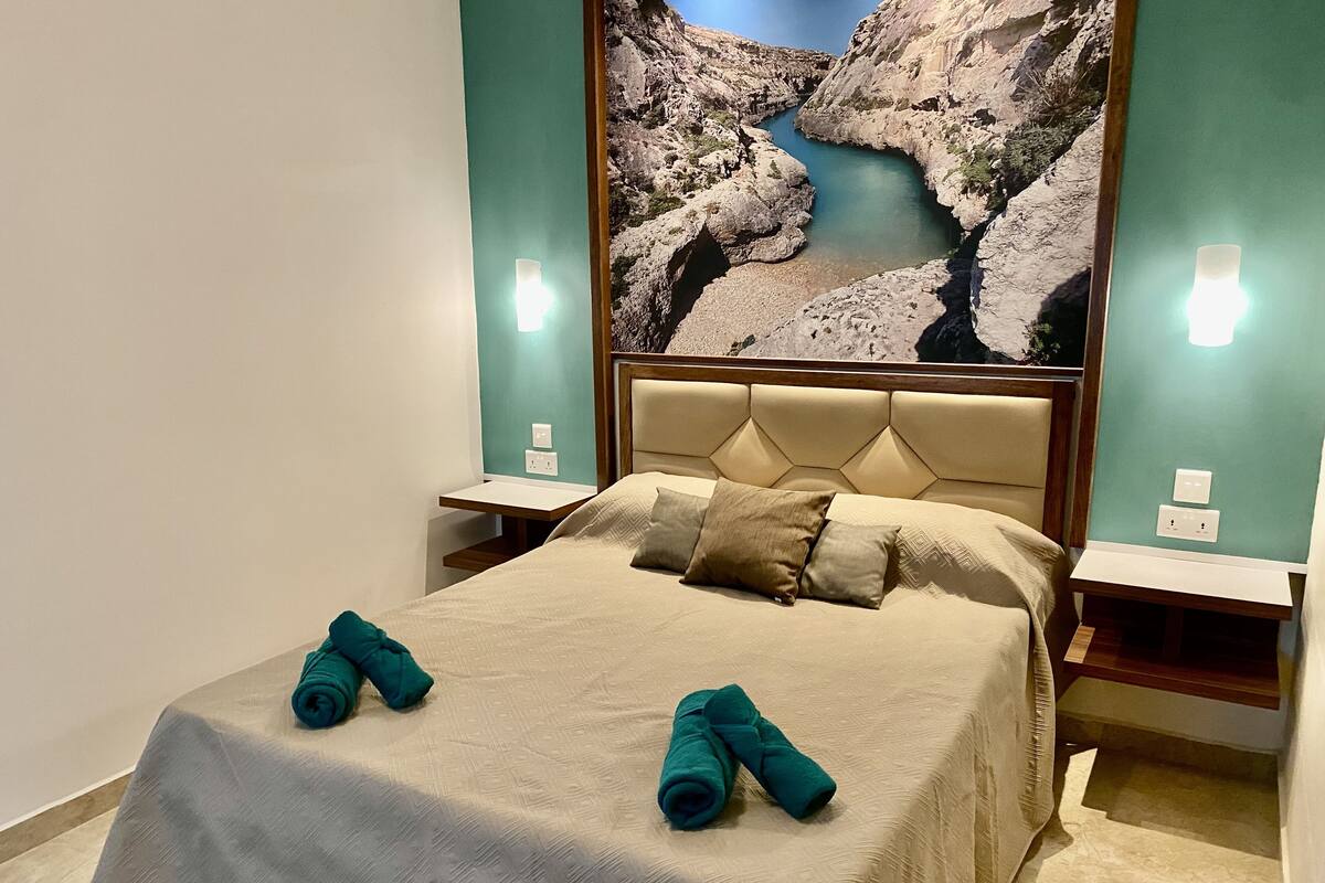 Habitación de hotel con cama, dos mesitas de noche y un cuadro colgado en la pared que representa un paisaje rocoso.