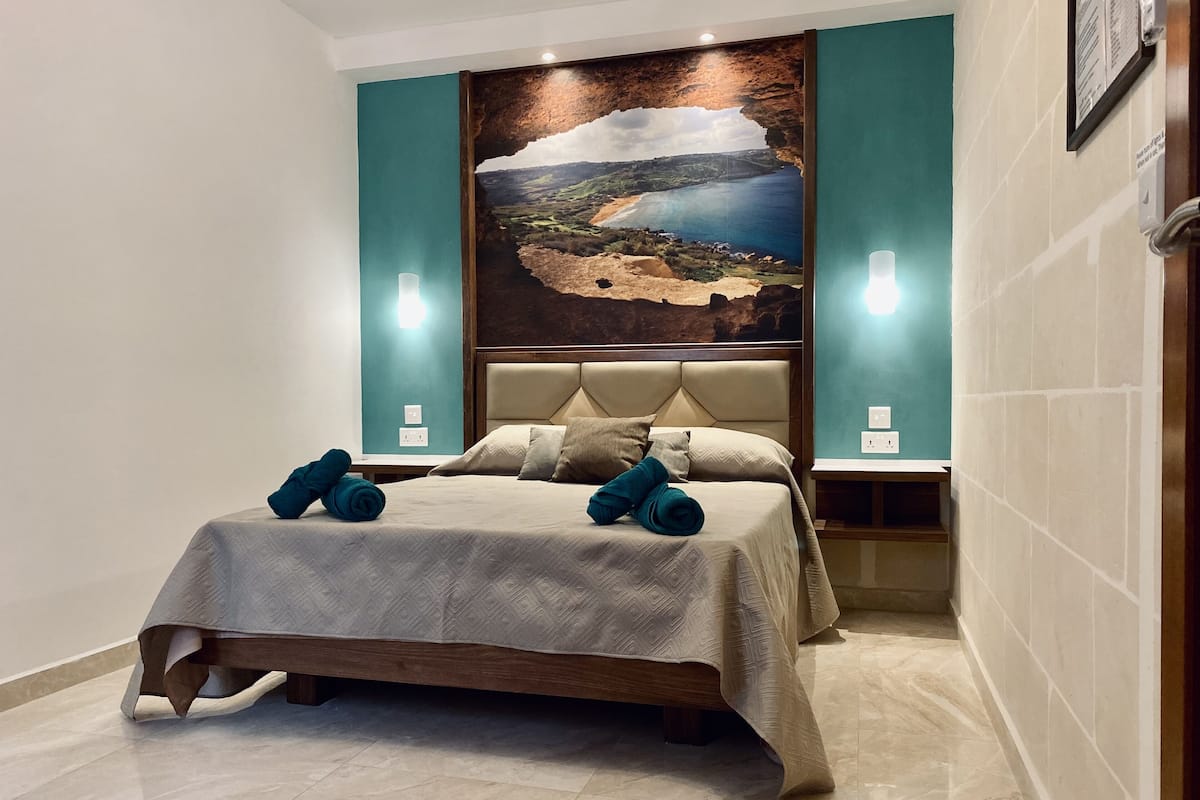 Una habitación de hotel con una cama grande, un cabecero con una pintura de paisaje y dos lámparas de noche.