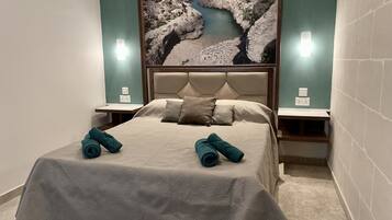 A neatly made bed with a beige bedspread and two rolled towels, flanked by nightstands with wall-mounted lamps, in a room with a large rock and river mural on the wall.