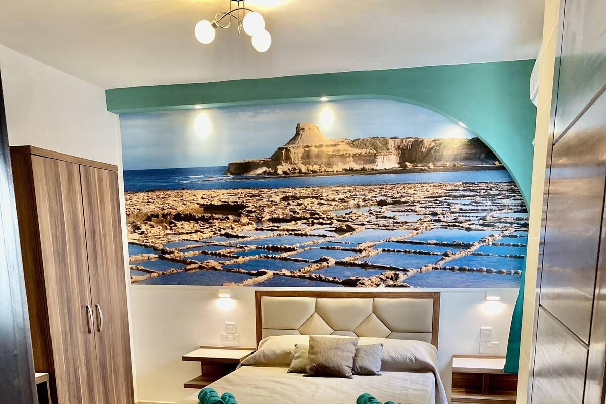 Un dormitorio con una cama grande, un armario de madera, una mesita de noche y un mural en la pared con un paisaje costero.