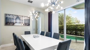 A modern dining room with a white table and black chairs, a large window with a view of a swimming pool, and a ceiling light fixture. - Lovely 5 Bed Family Pool Home in Reunion-7786LL (Kissimmee)