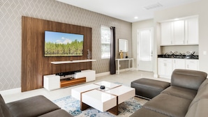 A modern living room with a flat-screen TV, a wooden entertainment unit, a sofa, a coffee table, and a kitchen area with white cabinets and a countertop. - Lovely 5 Bed Family Pool Home in Reunion-7786LL (Kissimmee)
