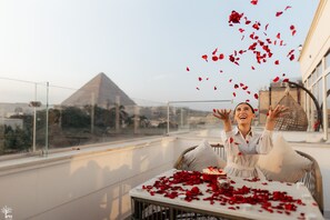 A balcony with a table covered in rose petals, a person sitting at the table, and a view of a pyramid in the background. - Tree Lounge Pyramids View INN, Sphinx Giza. (Giza)