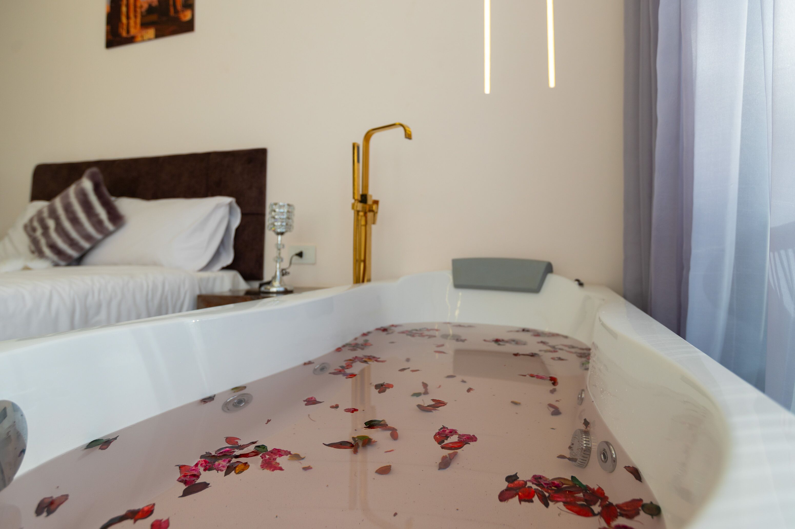 A hotel room with a bathtub filled with rose petals, a bed with a striped pillow, and a gold faucet.
