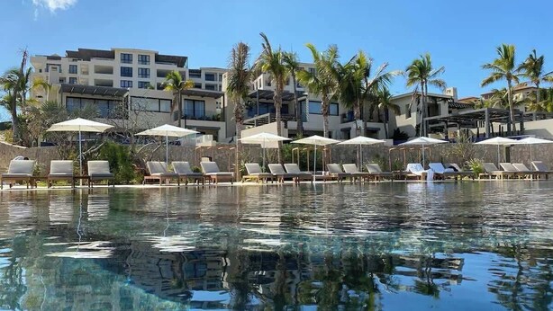 Una piscina cristalina con sillas de descanso y sombrillas, rodeada de palmeras y edificios modernos.