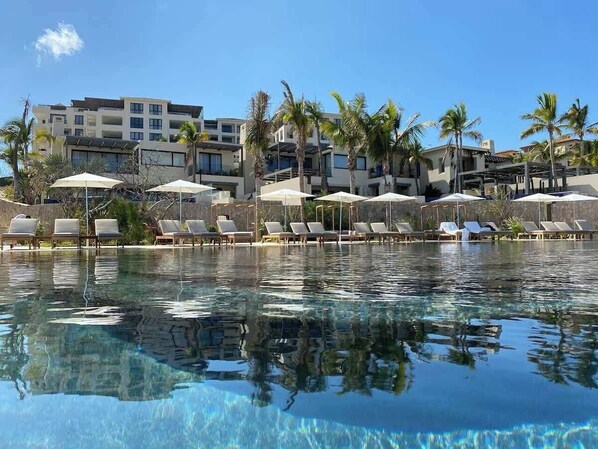 Ein klares Schwimmbecken mit Liegestühlen und Sonnenschirmen, umgeben von Palmen und modernen Gebäuden.