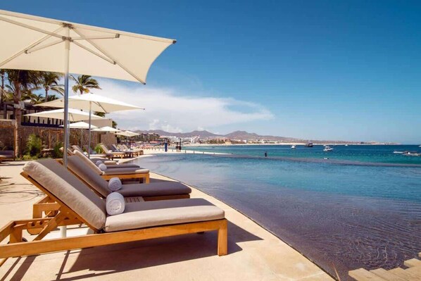 Um resort na praia com espreguiçadeiras e guarda-sóis, piscina e vista para o oceano e montanhas ao longe.