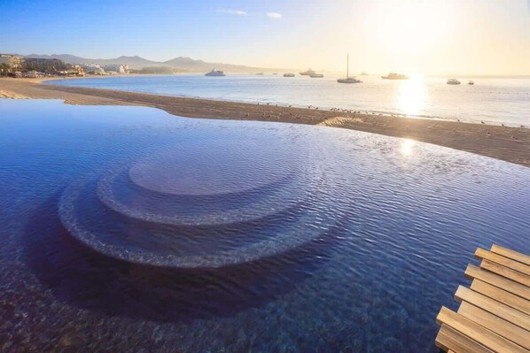 Ein klarer Blick auf einen Infinity-Pool mit einer Holzterrasse, der auf ein ruhiges Meer mit Booten und einer entfernten Küstenlinie blickt.