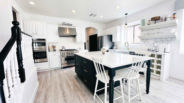 Una cocina moderna con armarios blancos, una isla central con taburetes de bar, electrodomésticos de acero inoxidable y un letrero decorativo sobre la estufa.
