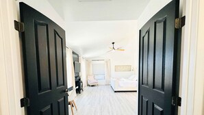 Un pasillo que conduce a un dormitorio con cama, una silla, televisor y ventilador de techo.