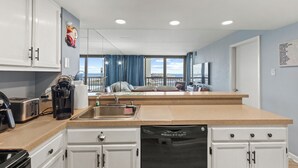 A modern kitchen with white cabinets, a dishwasher, a coffee maker, and a view of the city through large windows. - Sea Watch 505 by Seaside Vacations & Sales (Ocean City)