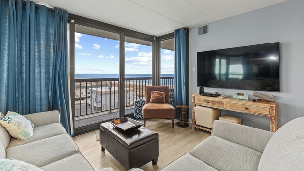 A living room with a large TV, a sofa, a chair, a desk, and a view of the beach. - Sea Watch 505 by Seaside Vacations & Sales (Ocean City)
