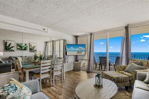 Un salón moderno con un televisor grande, suelo de madera y un balcón con vistas al mar.