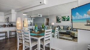Una cocina moderna con armarios blancos, un comedor con mesa de madera y sillas blancas, y una sala de estar con sofá y un cuadro grande con motivos playeros.
