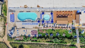 Vista aérea de un resort con una gran piscina, una piscina circular más pequeña y varias tumbonas.