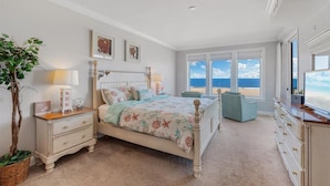 Ein Schlafzimmer mit einem Bett, Nachttischen, einem Schminktisch und einem großen Fenster mit Blick auf den Strand.