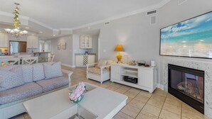 Ein großzügiges Wohnzimmer mit einer Couch, einem Couchtisch und einem Kamin. Im Hintergrund befindet sich eine Küche mit weißen Schränken und einem Essbereich.