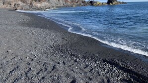 Una playa de guijarros oscuros, un acantilado rocoso y una formación rocosa destacada en el agua.