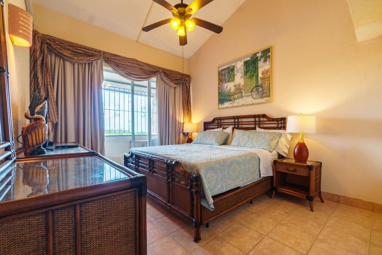 A bedroom with a wooden bed, a desk, a chair, a ceiling fan, and a painting on the wall.