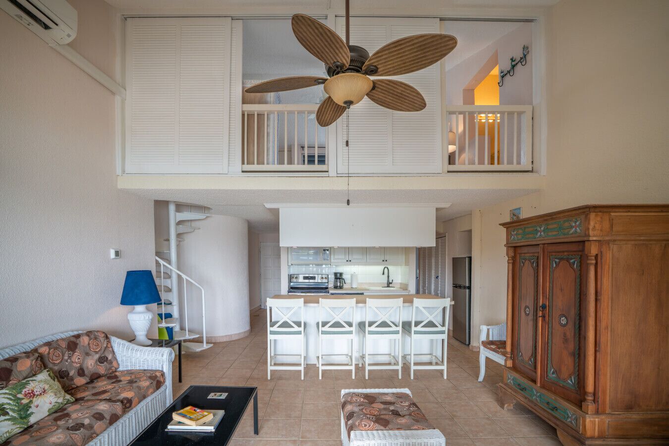 Un vaste espace de vie agrémenté d’un ventilateur de plafond, une cuisine avec des meubles blancs, une table à manger avec des chaises et un buffet en bois.