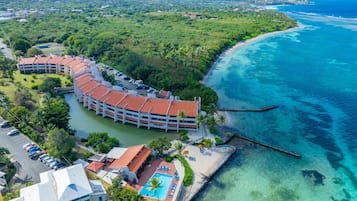 Vue aérienne d’une station balnéaire avec un grand bâtiment doté d’un toit incurvé, d’une piscine et d’une zone de plage.