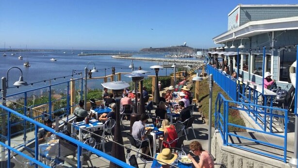 Zona de comedor al aire libre con mesas y sillas, con vista a un puerto deportivo lleno de barcos.