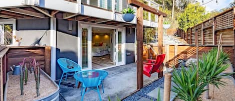 Une maison moderne dotée d’un patio comprenant une table et des chaises, un fauteuil suspendu et un petit jardin avec des cactus et des plantes.