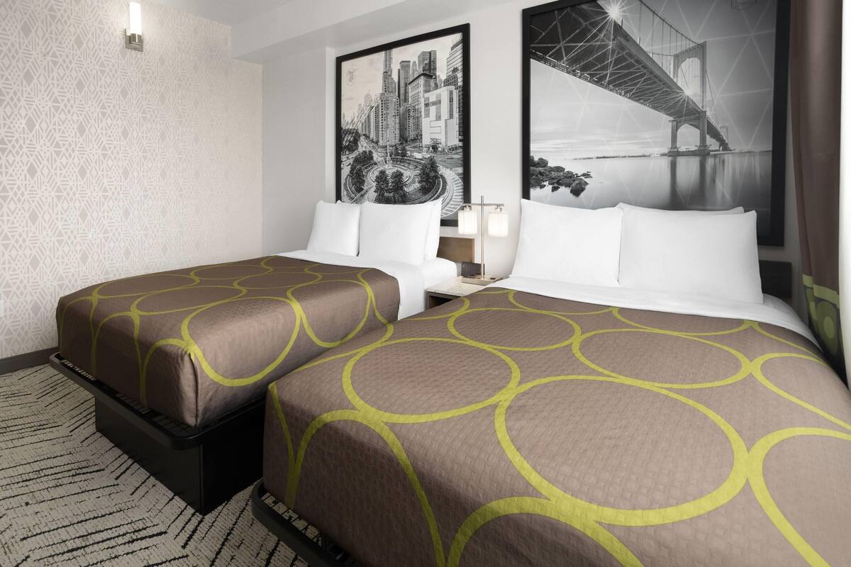 Habitación de hotel con dos camas, un papel tapiz estampado y dos cuadros con vistas urbanas.