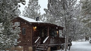 A wooden cabin with a porch, surrounded by snow-covered trees. - Western theme! | Amazing Spa, Fireplace and Deck! (Overgaard)