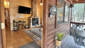 A cozy cabin interior with a stone fireplace, wooden walls, a mounted TV, and a porch with outdoor seating. - Western theme! | Amazing Spa, Fireplace and Deck! (Overgaard)