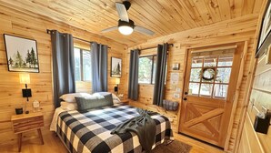 A cozy wooden cabin bedroom with a bed, two bedside tables, a ceiling fan, and a door with a window. - Juniper Grove | Loft & Personal Hot Tub! (Overgaard)