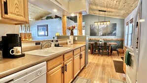 A modern kitchen with wooden cabinets, a built-in dishwasher, a coffee maker, and a dining area with a table and chairs. - The Prancing Pony | Hot Tub & Quiet Location! (Overgaard)