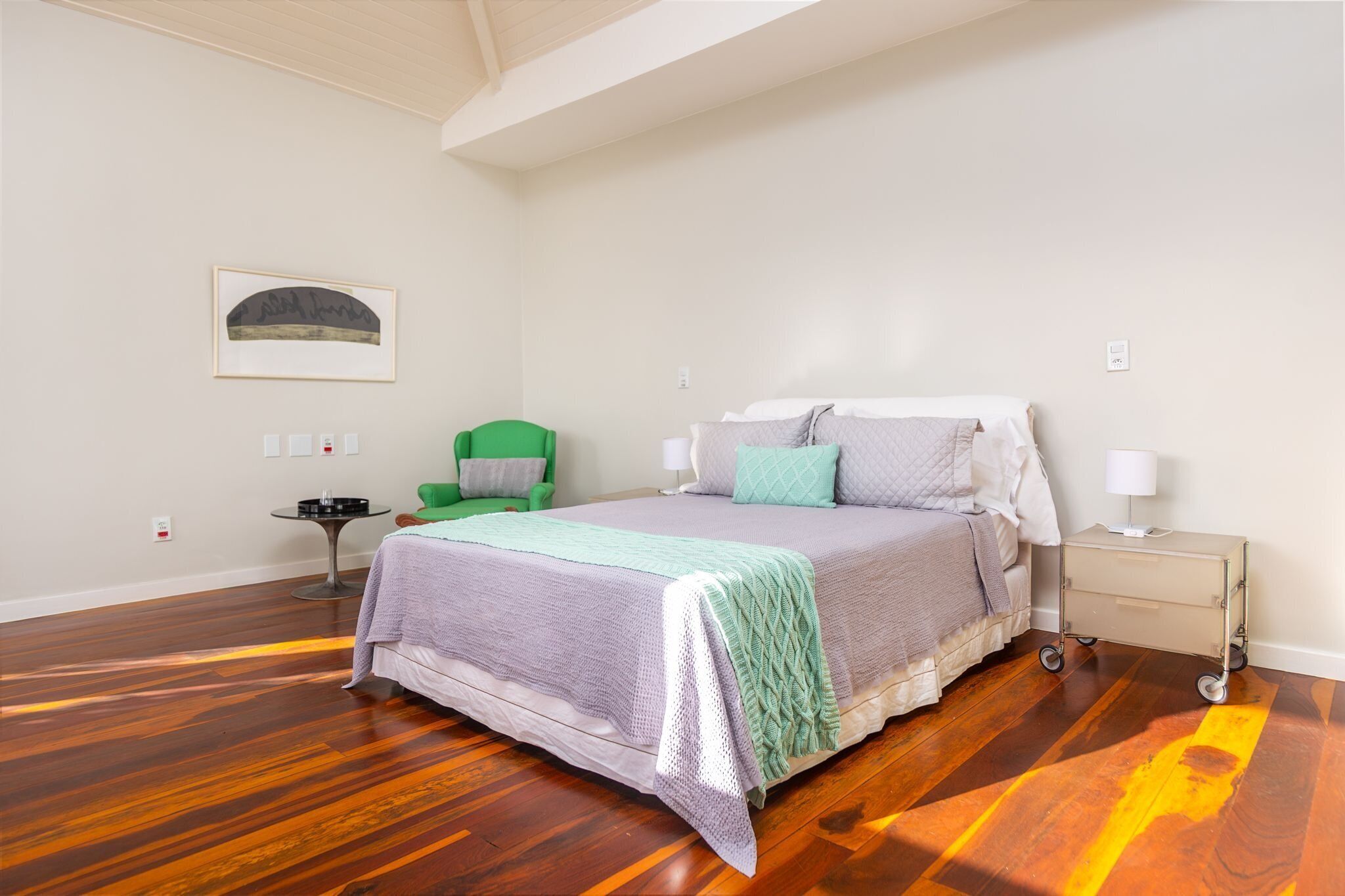 Quarto com cama, mesa de cabeceira, abajur, cadeira verde e quadro na parede.