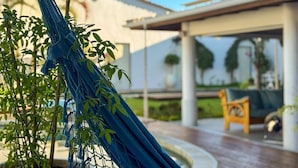 Un hamac attaché à un arbre, un espace patio avec une chaise en bois, et un bâtiment à la façade blanche doté d’une partie abritée.