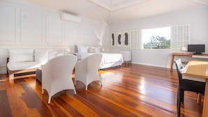 Une pièce spacieuse avec un plancher en bois, un lit recouvert de linge de lit blanc, deux chaises en osier blanches, une petite table et un téléviseur.