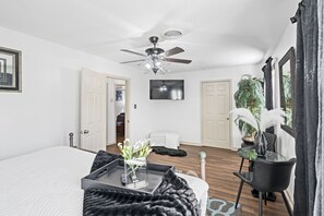 Un dormitorio con una cama grande, ventilador de techo, televisor, zona de comedor con mesa y sillas, y una planta en maceta.