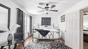 A bedroom with a bed, bedside tables, a ceiling fan, a window with curtains, and a door.