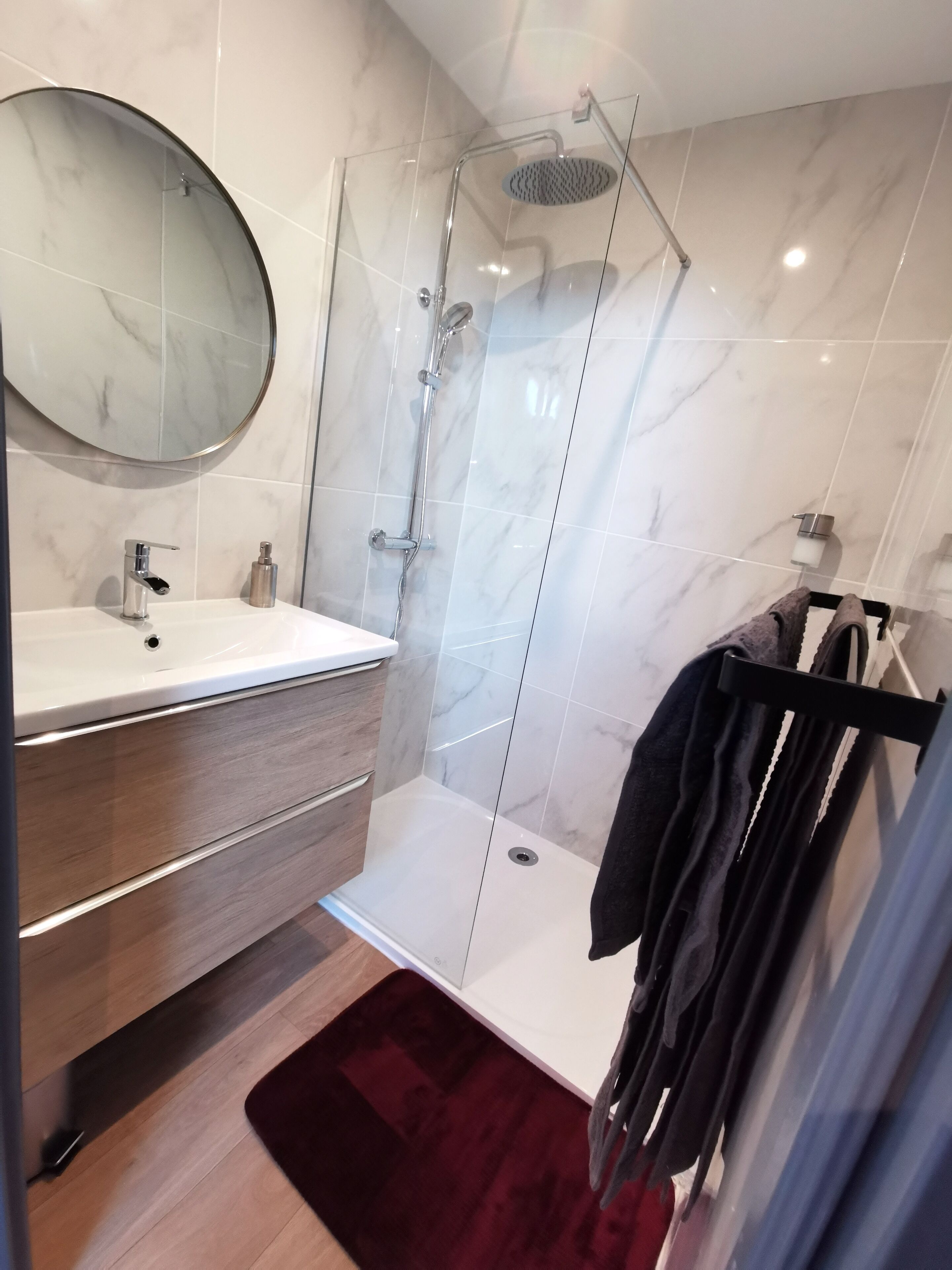 Un baño moderno con ducha de vidrio, un espejo redondo, un lavamanos blanco y un mueble de tocador de madera.