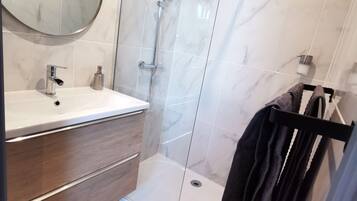 Un baño moderno con ducha de vidrio, un espejo redondo, un lavamanos blanco y un mueble de tocador de madera.