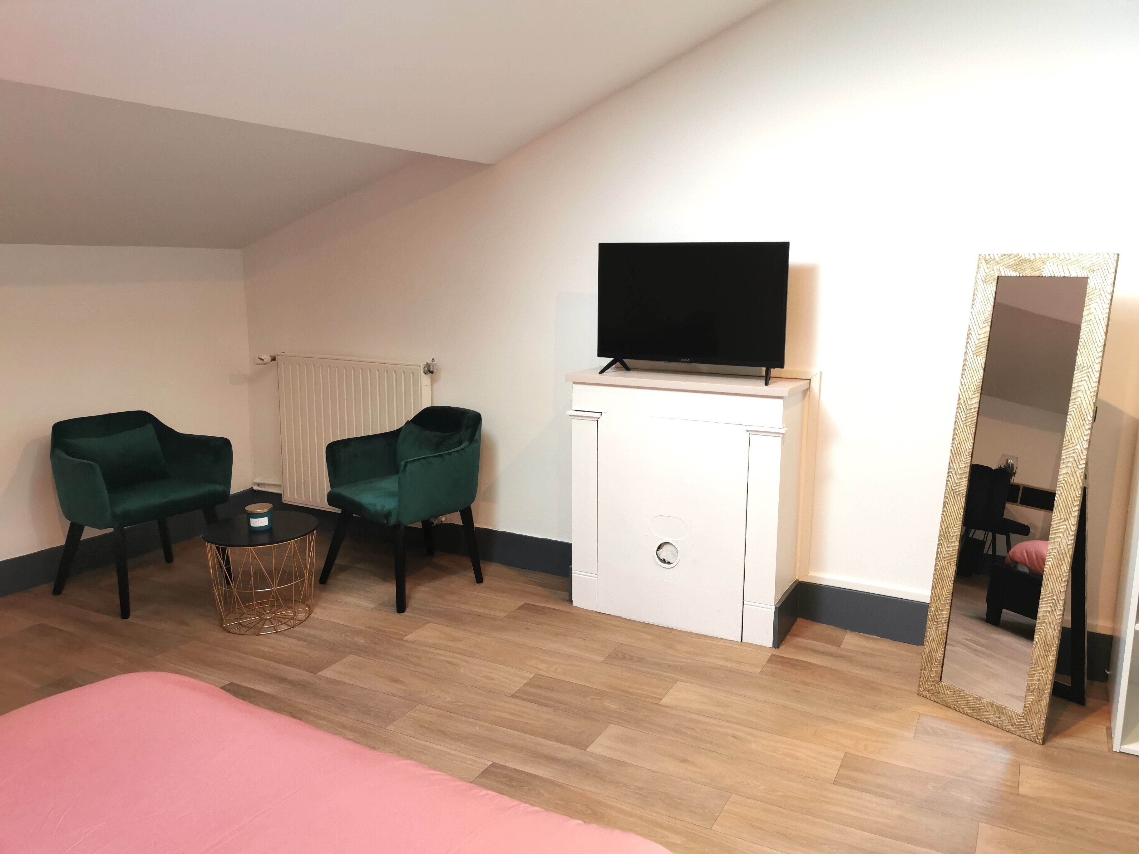A room with a bed, two green armchairs, a small table with a basket, a white cabinet with a TV, and a large mirror.
