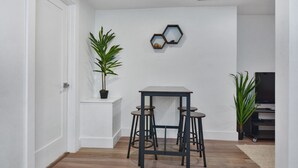 A small, modern room with a white door, a wooden floor, a small table with stools, a potted plant, and a hexagonal mirror on the wall. - Sleek Somerville Apartment near Encore! (Somerville)